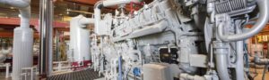 Large industrial machinery with multiple pipes, valves, and metal components—including essential Waukesha engine parts—is situated inside a factory setting with overhead beams and various equipment in the background.