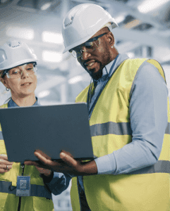 Two construction workers wearing hard hats, safety glasses, and yellow reflective vests look at a laptop together in an industrial setting, possibly consulting a heavy equipment parts supplier for their project needs.