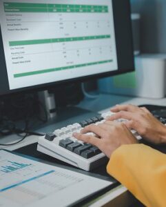 A person wearing a yellow sleeve types on a keyboard in front of a computer monitor displaying a spreadsheet; nearby, a printed chart with graphs suggests they’re analyzing data for a heavy equipment parts supplier.