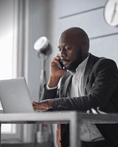 A man in a suit sits at a desk, talking on a smartphone and using a laptop—perhaps coordinating as a heavy equipment parts supplier. A large clock hangs on the wall behind him while natural light pours in through the window.