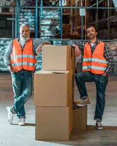Two men in orange safety vests stand in a warehouse, smiling and leaning on a stack of four large boxes. The background highlights shelves and packaging materials at a heavy equipment parts supplier facility.